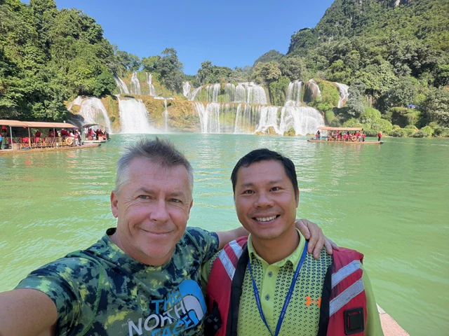 Two travellers on a boat in front of a wide waterfall in Vietnam