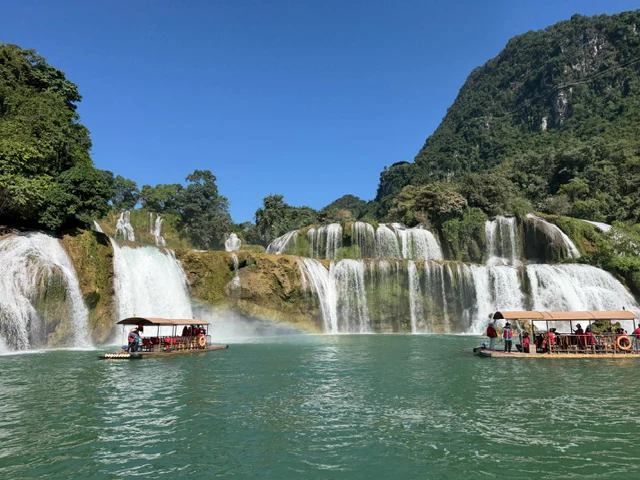 Covered passenger boats on turquoise water below a wide tiered waterfall in Vietnam